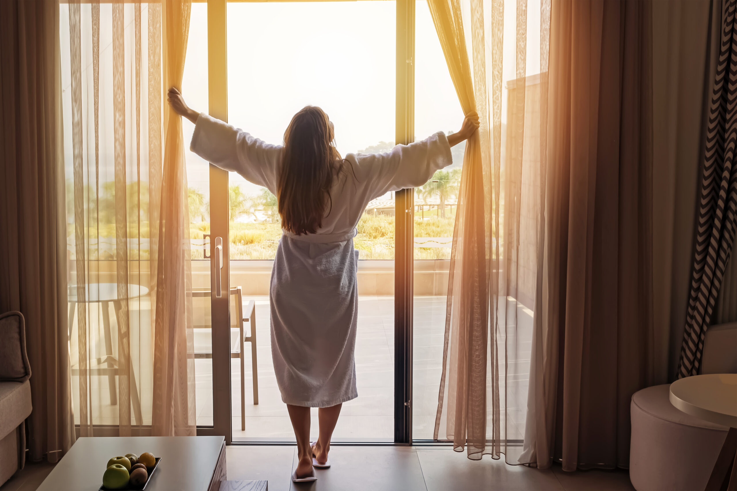 Young woman wearing white bathrobe opening curtains in luxury hotel room